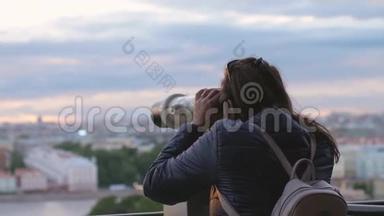 年轻的女游客带着背包，在日落时分用<strong>投币</strong>式双筒望远镜缓慢地观察城市